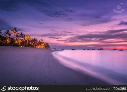 Sandy beach with palm trees at colorful sunset in summer. Tropical landscape with sea shore, blurred water, palms, boats and yachts in ocean, purple sky with clouds at night. Travel in exotic Africa