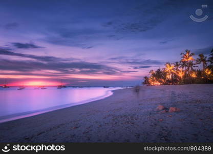 Sandy beach with palm trees at colorful sunset in summer. Tropical landscape with sea shore, blurred water, palms, boats and yachts in ocean, purple sky with clouds at night. Travel in exotic Africa