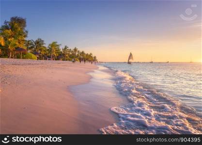 Sandy beach with sea waves, palm trees and walking people at colorful sunset in summer. Tropical landscape with blue sea, palms, boats and yachts in ocean, beautiful sky. Travel in exotic Africa