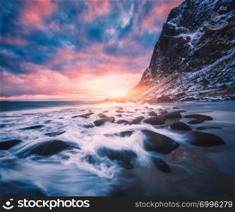 Sandy beach with stones in blurred water, blue sky with pink clouds and snowy mountains at sunset. Utakleiv beach, Lofoten islands, Norway. Winter landscape with sea, waves, rocks in the evening