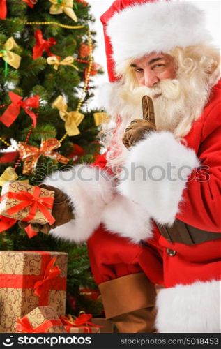 Santa Claus bringing gifts and putting under Christmas tree with silence gesture