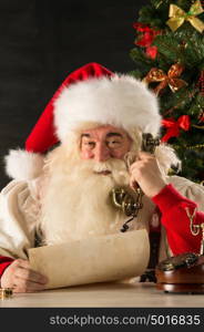 Santa Claus calling with vintage phone while reading an old roll letter. Sitting near Christmas tree at home