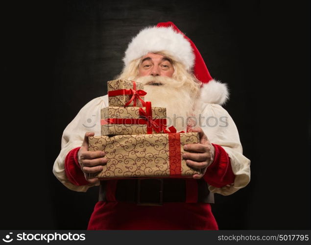Santa Claus carrying big stack of Christmas gifts against dark background