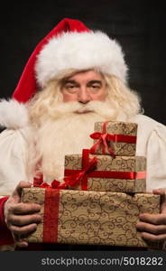 Santa Claus carrying big stack of Christmas gifts against dark background