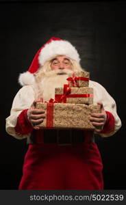 Santa Claus carrying big stack of Christmas gifts against dark background