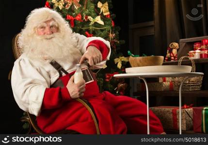 Santa Claus eating cookies with milk sitting near Christmas tree. Opening milk bottle