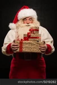 Santa Claus holding stack of Christmas gifts against dark background