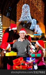 Santa Claus in male and female variants giving Christmas gifts in a shopping mall amid artificial snow covered fir trees and lights
