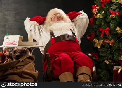 Santa Claus keeping his hands behind head while relaxing at home sitting in comfortable armchair near christmas tree drinking milk and eating fresh cookies. For Santa sin on table