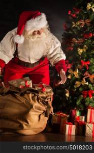 Santa Claus opening his sack and taking gifts under Christmas tree at night at living room at children's home