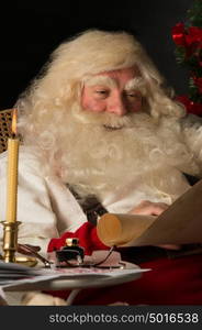 Santa Claus sitting at home and reading old paper roll to do list at night with candle light. Authentic vintage style portrait.