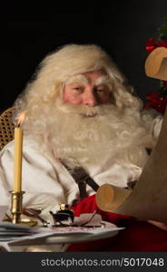 Santa Claus sitting at home and reading old paper roll to do list at night with candle light. Authentic vintage style portrait.