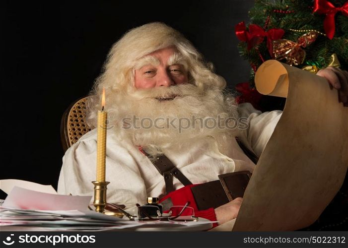 Santa Claus sitting at home and reading old paper roll to do list at night with candle light. Authentic vintage style portrait.