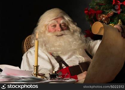 Santa Claus sitting at home and reading old paper roll to do list at night with candle light. Authentic vintage style portrait.