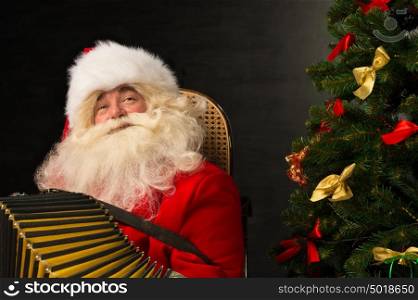 Santa Claus sitting in armchair near Christmas Tree at home and playing music on accordion