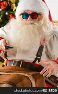 Santa Claus sitting in rocking chair near Christmas Tree at home and watching tv or home theater wearing 3d glasses and holding remote control
