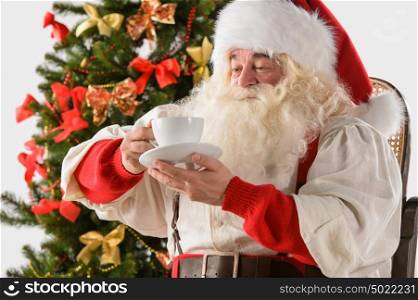 Santa Claus sitting in rocking chair near Christmas Tree at home and drinking hot tea or coffee