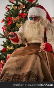 Santa Claus sitting in rocking chair near Christmas Tree at home and watching tv or home theater wearing 3d glasses and holding remote control