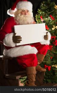 Santa Claus sitting indoors at dark room near Christmas tree and holding blank sign