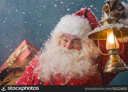 Santa Claus walking on the snow with his sack of lots of gifts and lights his way with vintage lantern. Winter night with snowfall