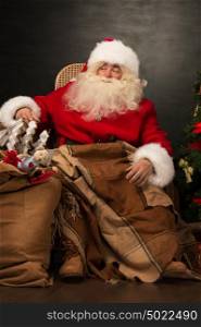 Santa Claus with a huge bag full of Christmas presents sitting in a comfortable chair near the Christmas tree at home