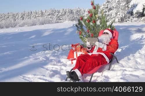 Santa Claus with gift bag using tablet computer in snow covered winter forest near Christmas tree