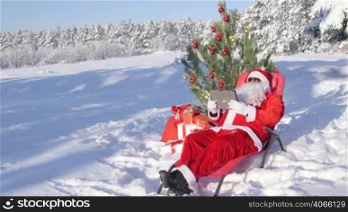 Santa Claus with gift bag using tablet computer in snow covered winter forest near Christmas tree