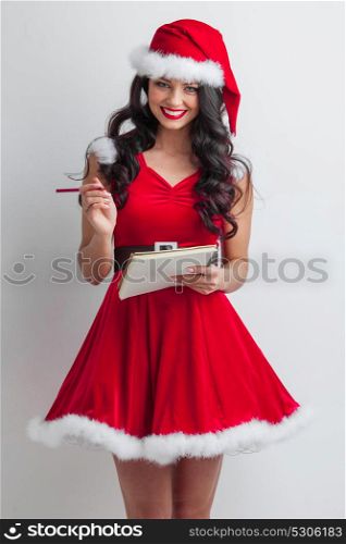 Santa girl with wish list. Pretty smiling pin-up Santa girl in red dress with wish list and pencil