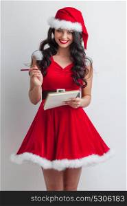 Santa girl with wish list. Pretty smiling pin-up Santa girl in red dress with wish list and pencil