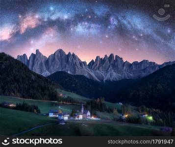 Santa Maddalena and acrhed Milky Way at night in summer in Italy. Starry sky with milky way arch over St. Magdalena and mountains. Village with houses, church, green meadows, trees, rocks. Space