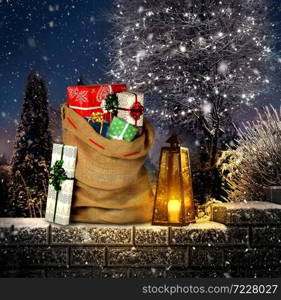 Santas present sack with gift boxes on winter outdoor garden settting with lantern and candle. Gingerbread cottage with Christmas tree