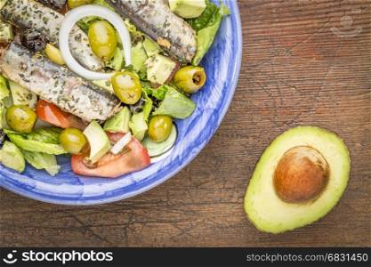 sardine salad (canned sardines, romaine lettuce, tomato, avocado, onion, olives) in bowl against grunge wood with a half of avocado
