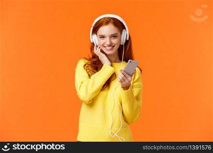 Sassy good-looking redhead female in yellow sweater, listen music white headphones, touch earphones and holding smartphone, listen music, enjoy good earbuds quality, smiling, orange background.. Sassy good-looking redhead female in yellow sweater, listen music white headphones, touch earphones and holding smartphone, listen music, enjoy good earbuds quality, smiling, orange background