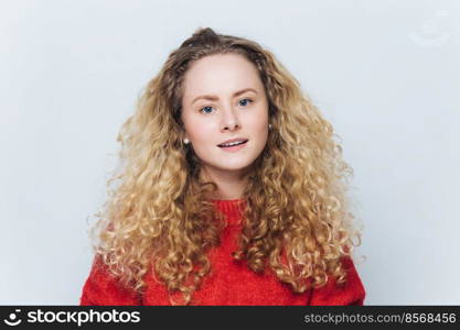 Satisfied adorable blue eyed female with curly light hair, wears red sweater, has curious expression, isolated over white background. Attractive woman has blonde hair, poses in studio alone.