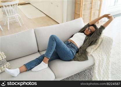 Satisfied african american young girl stretχng after day nap, lying on comfortab≤sofa in living room at home. Relaxed sere≠fema≤resting on cozy couch enjoying break on lazy weekend.. African american girl stretχng after day nap, lying on comfortab≤sofa in living room at home