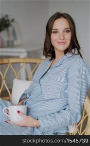 Satisfied beautiful woman with pleasant appearance, wears striped nightwear, drinks aromatic coffee, poses indoor. Relaxed housewife has drink at morning after awakening. People and rest concept