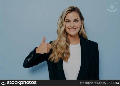 Satisfied blonde woman in formal outfit showing thumbs up gesture, giving positive feedback, demonstrating approving expression while looking at camera, standing alone next to blue background. Cheerful blonde girl showing thumbs up gesture