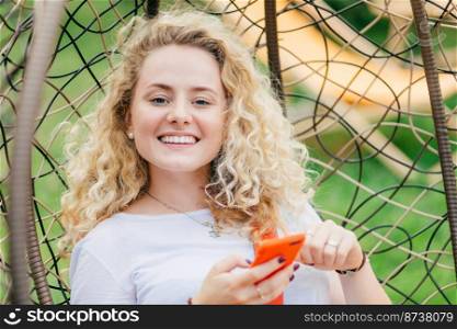Satisfied blonde young female with curly hair, has positive smile, uses modern cell phone for chatting with friends, can`t imagine her life without modern technology, looks positively at camera