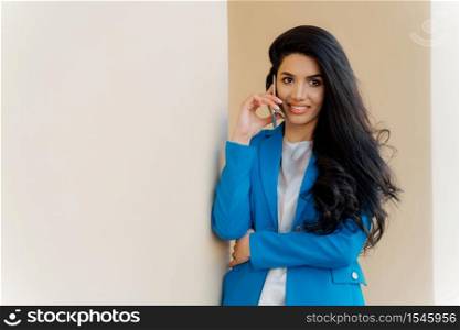 Satisfied brunette business lady calls on smartphone device, books tickets for trip abroad, makes positive conversation, dressed in formal elegant clothes, uses technology, poses near white wall.