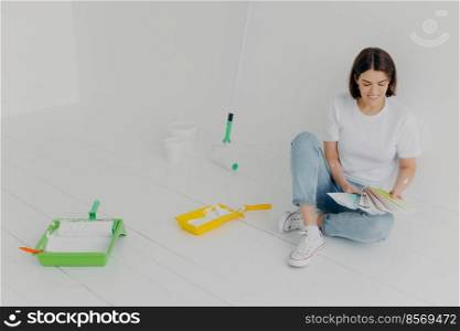 Satisfied brunette woman chooses color from s&les, going to refurbish room, sits on floor in casual clothes, surrounded with trays and paint rollers, busy with house renovation. DIY concept