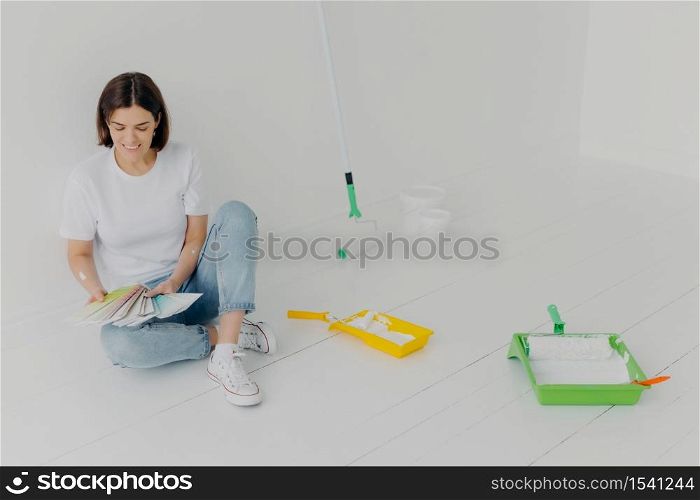 Satisfied brunette woman chooses color from samples, going to refurbish room, sits on floor in casual clothes, surrounded with trays and paint rollers, busy with house renovation. DIY concept