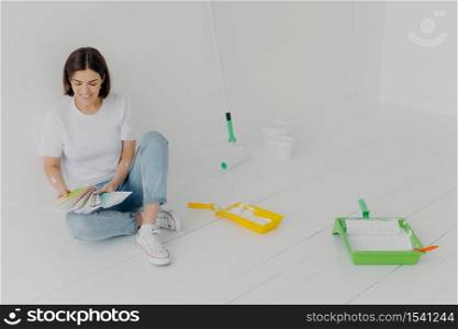 Satisfied brunette woman chooses color from samples, going to refurbish room, sits on floor in casual clothes, surrounded with trays and paint rollers, busy with house renovation. DIY concept