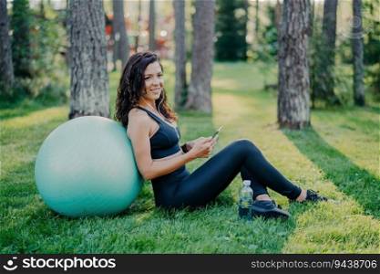 Satisfied brunette woman sits against big fitness ball, using her phone for music, on green grass in a sunny forest. Sporting concept, wearing sports clothes.