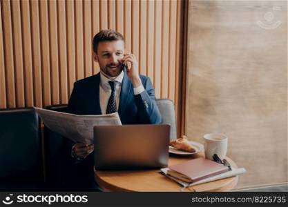 Satisfied business owner in blue suit talking on phone with newspaper in hand, sharing recent news with his partner and looking at laptop, notifying about further developments while working in cafe. Business owner talking on phone after reading news, sitting in cafe
