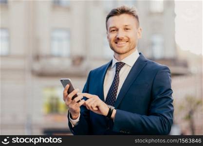 Satisfied businessman holds smart phone, reads email from investor, happy to recieve good news about business company, stands in urban territory, wears black suit and elegant tie. Technology concept