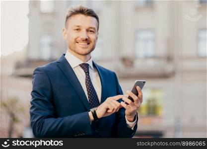Satisfied businessman holds smart phone, reads email from investor, happy to recieve good news about business company, stands in urban territory, wears black suit and elegant tie. Technology concept
