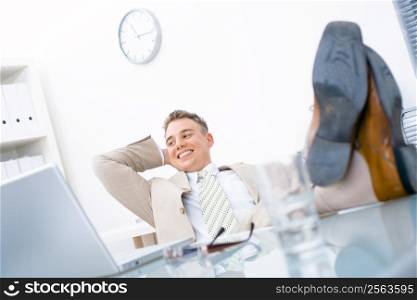 Satisfied businessman sitting by desk at office, feet on table, smiling.