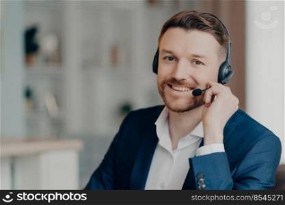 Satisfied cheerful bearded male office worker in suit consulting clients while sitting and using headset at home or office, selective focus on smiling man. Job and occupation concept. Happy young businessman wearing headphones with mic talking with clients indoors