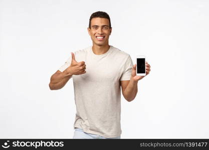 Satisfied, cheerful good-looking sexy young man in t-shirt, showing thumbs-up in approval or like gesture, smiling nod in agreement, recommend app, holding smartphone, introduce gadget.. Satisfied, cheerful good-looking sexy young man in t-shirt, showing thumbs-up in approval or like gesture, smiling nod in agreement, recommend app, holding smartphone, introduce gadget