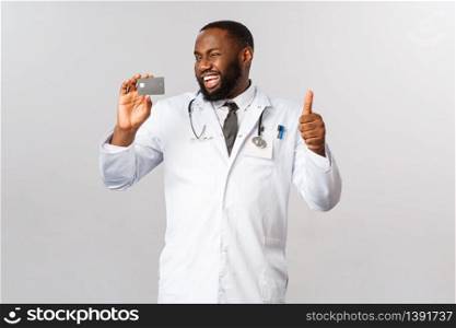 Satisfied, cheerful handsome african-american doctor in white coat, wink and show thumbs-up recommend credit card payment, contactless purchase during pandemic, new bank service.. Satisfied, cheerful handsome african-american doctor in white coat, wink and show thumbs-up recommend credit card payment, contactless purchase during pandemic, new bank service